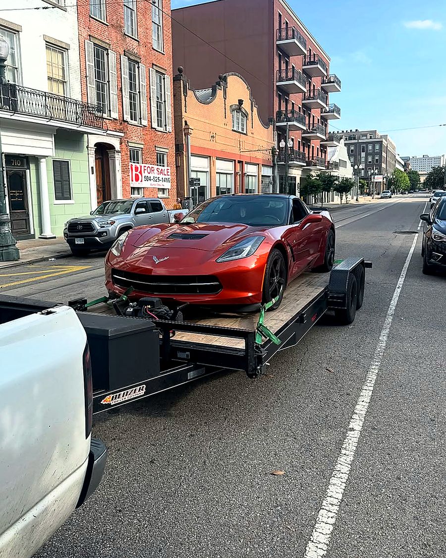 A red Chevrolet Corvette Stingray strapped to Rik's Towing flatbed trailer on a downtown New Orleans street.